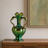 A green iridescent glass vase with a flared, leaf-like top and two curved handles, displayed on a wooden console table against a beige wall with framed artwork.