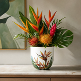 A vibrant tropical floral arrangement in a white ceramic pot with botanical illustrations, displayed on a marble countertop against a neutral beige wall.