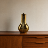 A polished eosin green metallic vase with a flared rim and vertical ribbed texture, displayed on a mid-century modern wooden sideboard against a neutral beige wall.