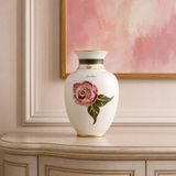 A white ceramic vase with a pink rose and green leaves, accented with gold trim, displayed on a beige console table against a pink wall with a framed painting.