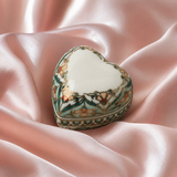 A heart-shaped porcelain keepsake box with a glossy white interior and an ornate floral pattern in teal, gold, and brown, resting on draped pink satin fabric.