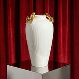 White vase with gold accents on a reflective surface in front of red curtains