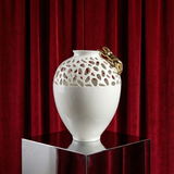 White decorative vase with gold accents on a reflective surface in front of red curtains