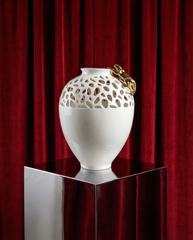 White decorative vase with gold accents on a reflective surface in front of red curtains