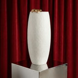 White textured vase with gold accents on a reflective surface in front of red curtains