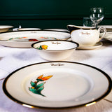 Dinnerware set with floral designs on a tablecloth, framed artwork in the background.
