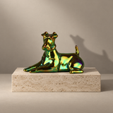 A glossy iridescent dog sculpture in shifting green, gold, and teal tones, posed lying down on a rectangular beige stone pedestal against a warm beige studio background.