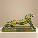 A polished green-gold metallic sculpture of a crouching jaguar on a rectangular base, displayed against a plain warm beige background.