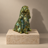 A glossy, metallic Cocker Spaniel dog sculpture in green and gold sits on a rectangular beige stone pedestal against a warm beige studio background.
