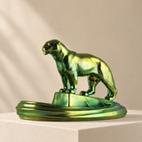 A metallic green panther figurine standing on a matching circular base, displayed on a white pedestal against a warm beige background with soft diagonal shadows.