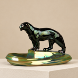 A glossy black panther figurine standing on a reflective iridescent gold and green base, displayed on a white pedestal against a warm beige background.