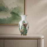 A white porcelain vase with a gold base and neck, decorated with a colorful tropical bird-of-paradise and bamboo pattern, displayed on a light beige console table against a beige wall with a framed green painting.