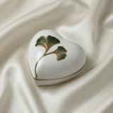 Heart-shaped white container with green leaf design on a beige fabric background