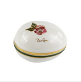 A white porcelain keepsake box with a lid decorated with a pink rose and green leaves, featuring a gold band around the base and gold script branding on top.