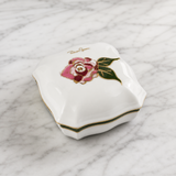 A white porcelain keepsake box with a lid, decorated with a pink rose, green leaves, and gold trim, resting on a white marble surface.
