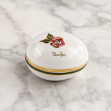 A white porcelain box with a lid, decorated with a pink rose, green leaves, and gold bands, resting on a white and gray marble surface.