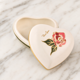 A heart-shaped ceramic keepsake box with a cream-colored exterior, gold trim, and a painted pink rose with green leaves on the lid, shown open on a white marble surface.