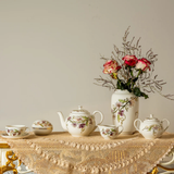 Tea set with floral patterns on a tablecloth and vase with flowers against a plain wall.