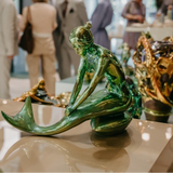 Green mermaid sculpture on a reflective surface with blurred people in the background