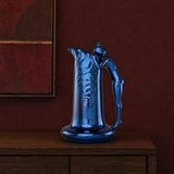 A glossy metallic blue pitcher with a sculptural handle, displayed on a dark wood cabinet against a deep red wall.