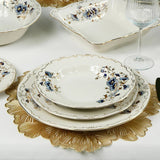 Dining table set with floral-patterned ceramic plates, gold-rimmed dishes, and glasses on a white tablecloth.