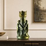 A decorative green and gold sculptural vase or candle holder with two classical figures and a flame-like top, displayed on a dark wood console in an elegant interior setting.